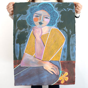 A woman holds a colorful painting on paper of an abstracted woman with blue hair, a yellow shirt and blue pants with tree shapes surrounding her, in front of  white wall. 