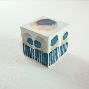 A white wooden cube with overlapping painted blue and beige shapes rests on a white surface. 