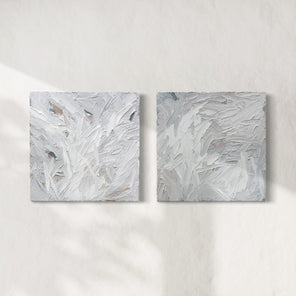 A pair of thickly textured paintings in grey, white, blue grey and plum grey by Teodora Guererra hanging on a white wall.