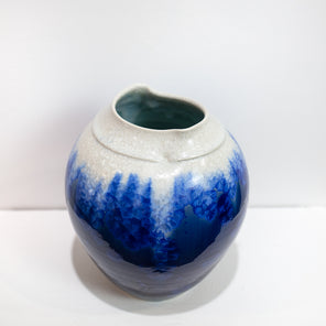 A blue and white glazed ceramic vase with a wide neck rests on a white surface in front of a white wall.