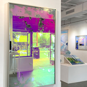 A pink fine art mirror hangs on a white column in an art gallery with paintings behind it. 